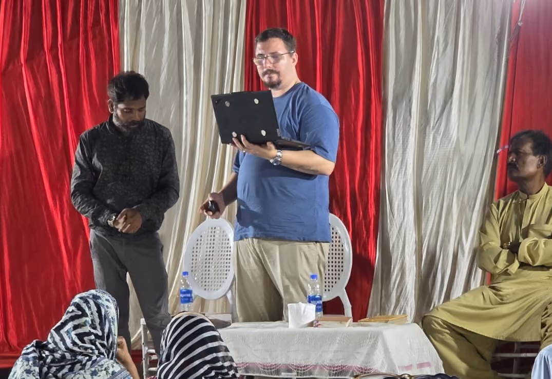 Sean teaching from a laptop to a seated gathering under red and white canopies, the listeners arms-folded and attentive.