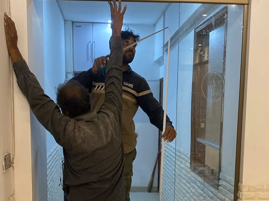 Two workmen fitting a glass door to the school premises, one steadying the frame overhead while the other measures behind the glass.