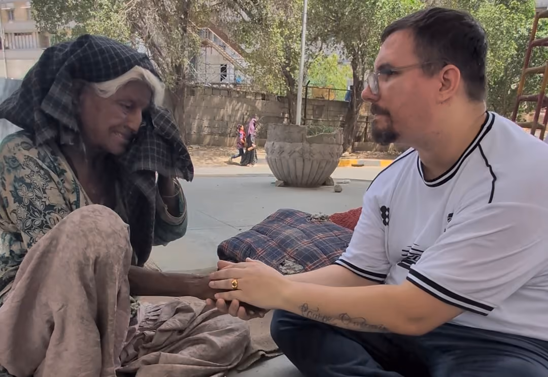 Two men sitting on the pavement facing each other, hands clasped between them, with tea and food set down alongside.