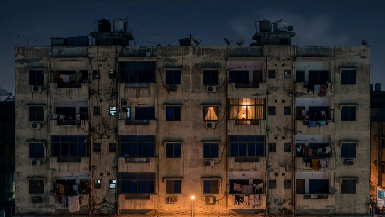 A Karachi apartment block at night, every window dark except one, lit from inside by a lamp.