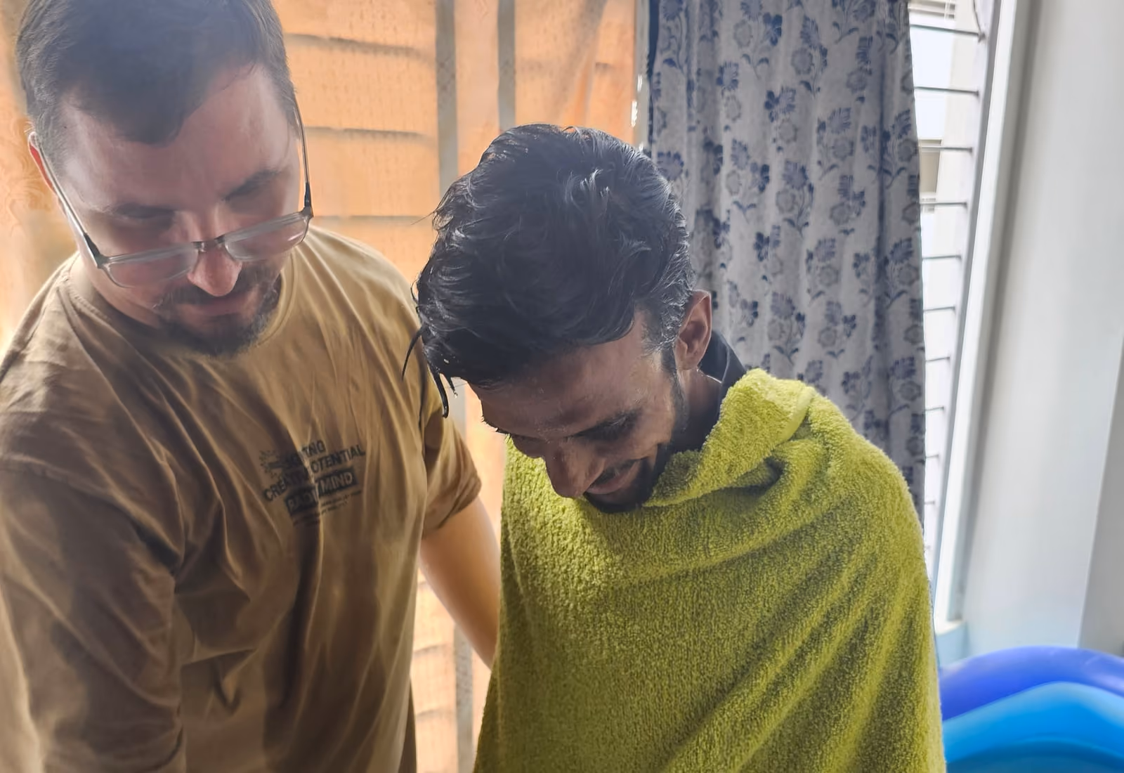 A young man wrapped in a towel in the moments after his baptism, steadied by the believer who baptised him.